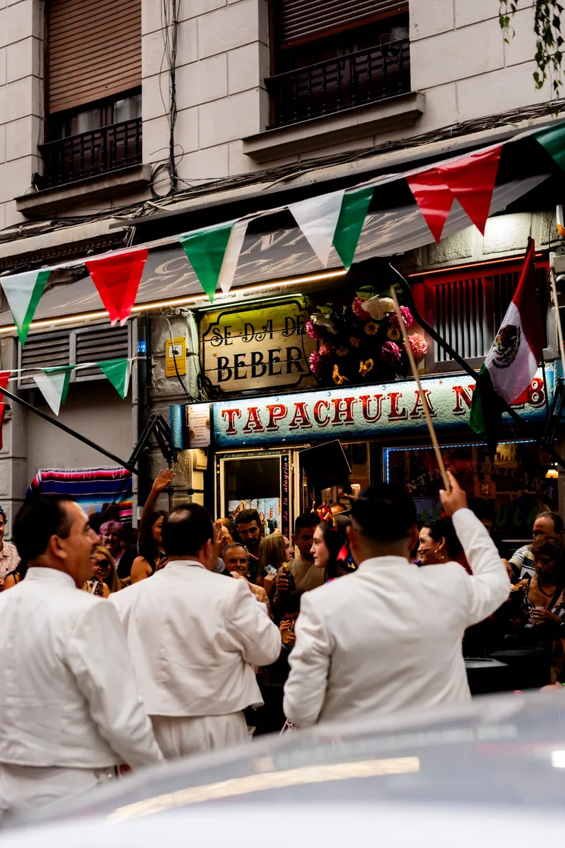 Mariachis vestidos de blanco y la calle llena ante la fachada de Cantina Tapachula, con banderas de México y papel picado